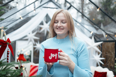 Smiling blonde Caucasian woman in light blue sweater holding a red mug with the word Joy near a...