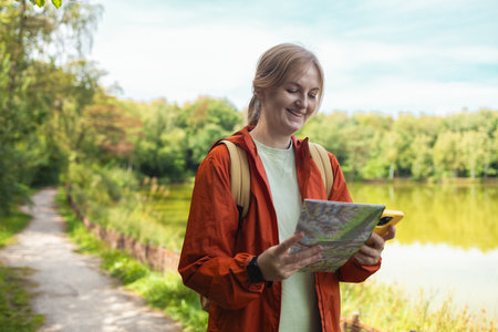 Happy young woman with a backpack wearing an orange jacket holding a paper map and smartphone while...