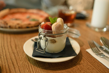 Delicious layered dessert with fresh strawberries and cream served in glass jar on wooden table in cozy cafe setting.