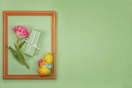 Greeting spring card for Easter, Mother's Day. Beautiful spring composition. Wooden frame of a lively terry tulip, packed in green paper, a gift and a figurine of a chicken with an Easter eggの写真素材