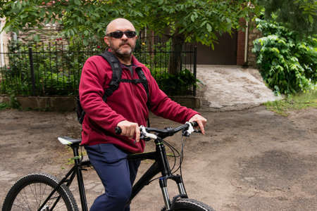 A senior man with a gray beard and a bald head in glasses and a red sweater with a hood on a snake sits on his mountain bike. Healthy sport and lifestyle in old age. Active life after isolationの写真素材