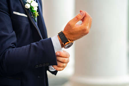 The groom in an expensive classic blue suit with a bouquet on the lapel adjusts the cufflink on the shirt cuff. Hands of a man in a suit with an expensive watch and a gold ring. Holiday. Valentine's Day. Weddingの写真素材