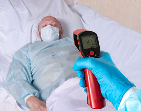 A doctor's hand in a glove holds an infrared thermometer and measures the temperature of the patient. Remote measurement of high fever for a coronovirus patient during a pandemic. The patient is blurred.の写真素材