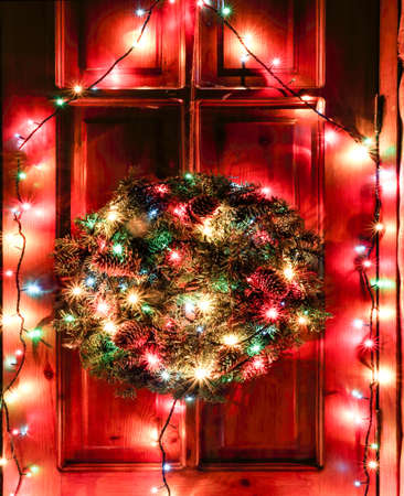 A wreath of Christmas tree branches with cones hanging on the front door. Night decoration of the door with a bright luminous garland, a wreath of fir branches and toys. Glowing lights on New Year's Eve.の写真素材