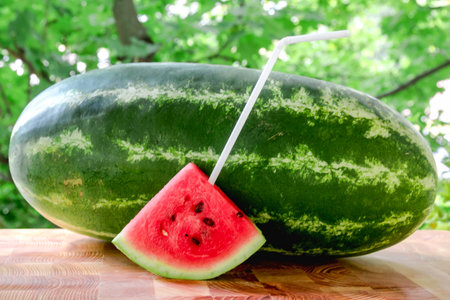 A triangular piece of a ripe watermelon with a straw stands in front of a long striped watermelon....