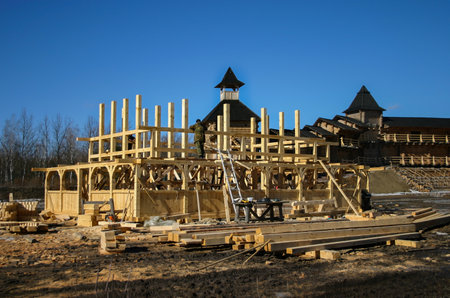 Construction of a traditional wooden house of uneven bars, logs and carved elements against the background of an ancient wooden fortress with towers. Construction site and tools. In the winter. blurryのeditorial素材
