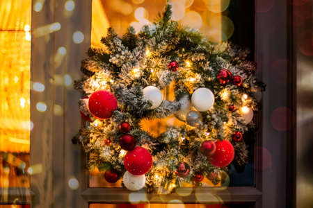 Christmas wreath made of fir branches with round balls of red, white and golden color and luminous garlands on the door. Decorating the door with a beautiful wreath for the New Year and holiday bokeh lightsの写真素材