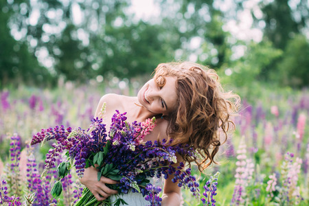 Beautiful young girl in a white dress holding a bouquet of lupine at sunset on the field. The toning.  The concept of nature and romance.の写真素材
