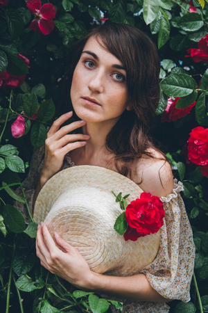 Girl with freckles in the hat stands against a background bushes with red roses. The toning. Selective focus.の写真素材