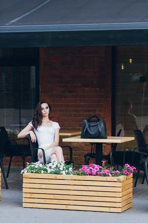 Young beautiful brunette girl with long hair in elegant white dress. Woman walking through the streets of the city in the evening at sunset.の写真素材