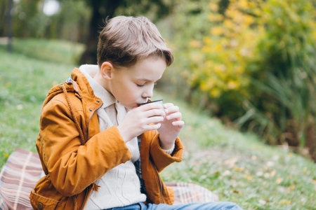 Cute boy walks and poses in a colorful autumn Park. The boy drinks hot tea from a thermos sitting on...