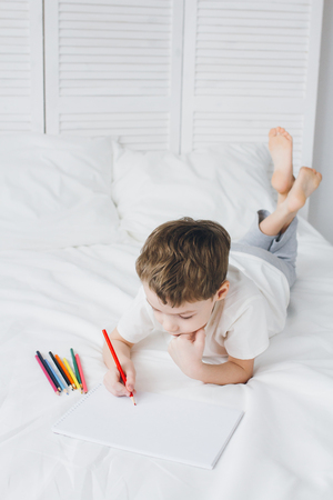 Cute boy draws with colorful pencils sitting on the bed with white linensの写真素材