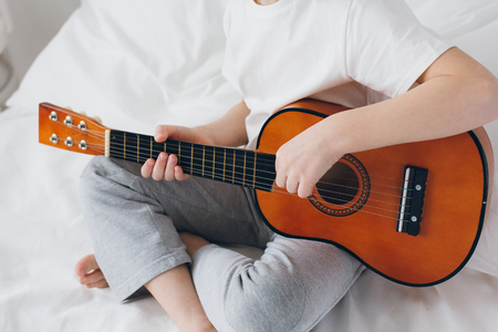 Cute boy playing a small guitar sitting on the bed with white linens. Selective focus.の写真素材