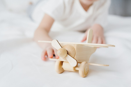 Cute boy playing with wooden airplane sitting on the bed with white linens. Selective focus.の写真素材