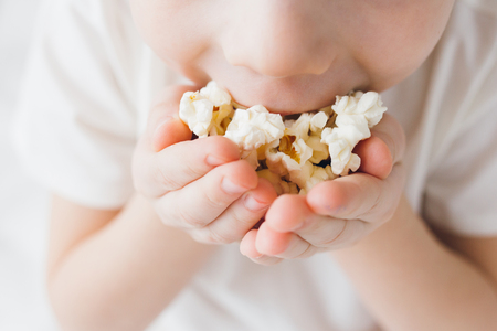 Cute boy eating popcorn sitting in bed with white linensの写真素材