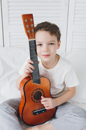 Cute boy playing a small guitar sitting on the bed with white linens. Selective focus.の写真素材