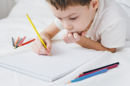 Cute boy draws with colorful pencils sitting on the bed with white linensの写真素材