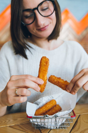 Young pretty brunette woman eats fried cheese sticks from a decorated iron basket in a cafe. Fast...