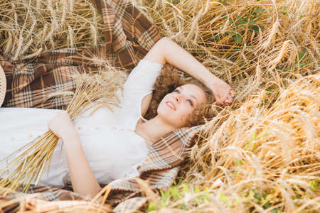 Young beautiful girl with long curly hair poses in a wheat field in the summer at sunset. The girl is lying on a blanket in the wheat. Toning.の写真素材