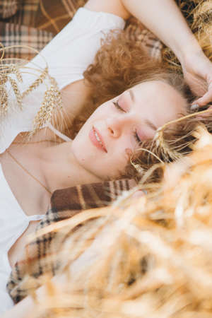 Young beautiful girl with long curly hair poses in a wheat field in the summer at sunset. The girl is lying on a blanket in the wheat. Toning.の写真素材