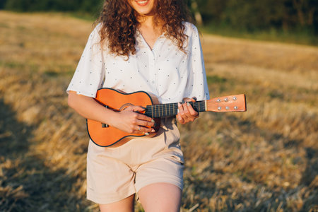 Young beautiful girl with long curly hair in shorts, a hat with a guitar poses in a wheat field in...