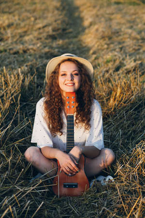 Young beautiful girl with long curly hair in shorts, a hat with a guitar poses in a wheat field in the summer at sunset. Toning.の写真素材