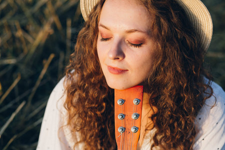 Young beautiful girl with long curly hair in shorts, a hat with a guitar poses in a wheat field in the summer at sunset. Toning.の写真素材