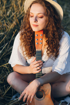 Young beautiful girl with long curly hair in shorts, a hat with a guitar poses in a wheat field in...