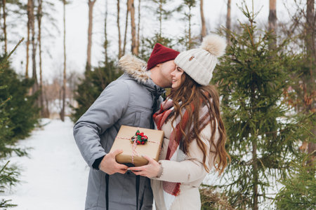 Young beautiful couple hugs and kisses in a winter coniferous forest, give each other boxes with gifts. A park with Christmas trees on the background. Christmas mood. Tinting.の写真素材