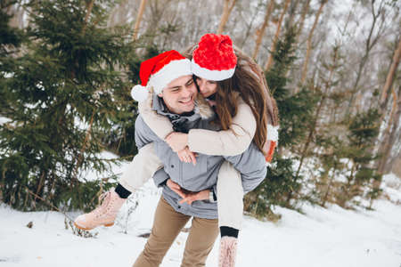 Young beautiful couple in a Santa hat and hugging and fooling around in a winter coniferous forest. Park with fir trees on the background. Christmas mood. Tinting.の写真素材