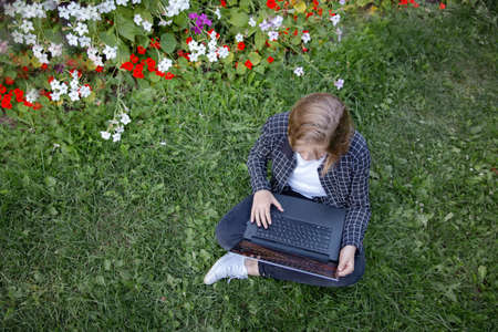 A teenage girl is working at a laptop, sitting in the garden on the grass among flowers and looking at the monitor. Teenage life, the concept of outdoor recreation. Top viewの写真素材