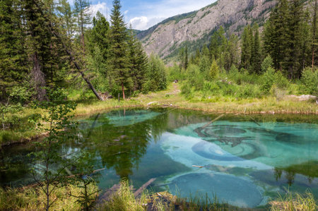 Beautiful geyser lake with clear turquoise water on a background of forest, mountains and blue sky with cloudsの写真素材