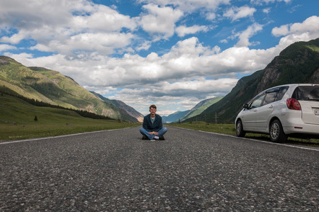 Smiling young man traveling by car in the mountains, sitting on the road.の写真素材