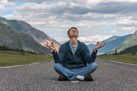 Young man sitting on the road among the mountains and meditating in the lotus position with legs crossed and eyes closedの写真素材