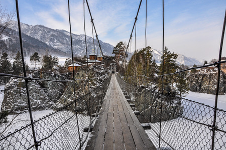 Suspension footbridge to the church across the frozen river in winter mountainsの写真素材