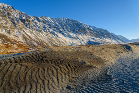 The picturesque mountain autumn landscape with snow covered mountains, frozen lake, sand dunes with sparse vegetation against the blue sky on a sunny dayの写真素材