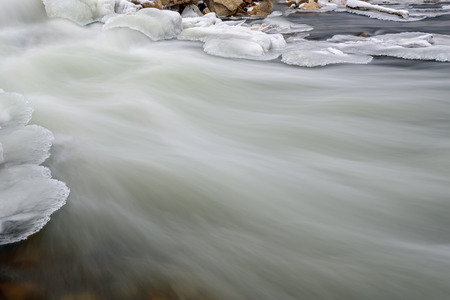 The picturesque view of the fast mountain river flowing among stones with banks of snow and ice. Shot with long exposure that provides silky water effect.の写真素材