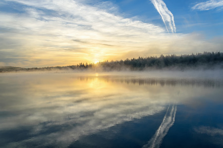 Colorful bright golden sunrise with fog on the lake with beautiful reflections of the clouds, the sun and the trees in the water against a blue skyの写真素材