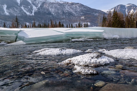 Scenic spring view on the fast mountain river flowing among stones with the banks of the ice and snow on background of mountains, forest and blue skyの写真素材