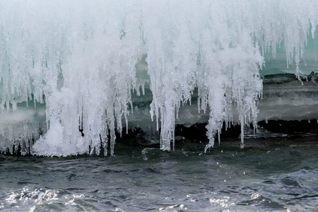Beautiful patterns of icicles of different shapes and sizes, turquoise layered ice and snow on the background of the rapid flow of water in the riverの写真素材