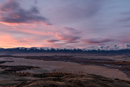 Scenic autumn landscape with a winding river and forest in the valley, snow mountains covered with snow and clouds in the pink light of the sun at dawnの写真素材