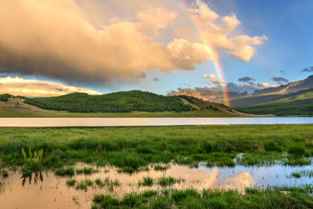 Bright colorful rainbow over the mountains, lake and forest after the rain against the blue sky with clouds in summerの写真素材