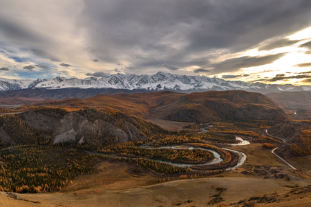Amazing autumn top view on the of the valley with a winding river, forest, golden trees mountains covered with snow on the background of the overcast sky and clouds at sunsetの写真素材
