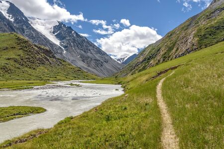 Amazing summer landscape with a path along the winding river along a green valley to the snowy mountains and the glacier against the blue sky with clouds. Altai, Russia.の写真素材