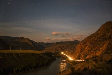 Night landscape with a winding road in the mountains, traces from the headlights of cars, a river, clouds and stars in the form of tracks in the skyの写真素材