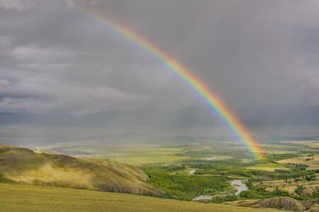 Amazing bright colorful rainbow over the mountains, a valley with a winding river and forest against a stormy sky with clouds and rain. Altai, Russia.