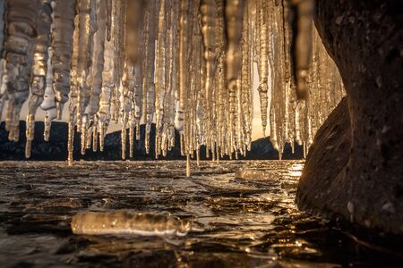 Amazing view of the shiny frozen ice floes on the lake, mountains and sun reflections through a curtain of icicles at sunset in winter. Teletskoye Lake, Altai, Russiaの写真素材