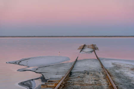 Delicate pink sunrise over a salt lake, rails going into the water and salt on the shore in the foreground. Lake Bursol, Altai, Russia.の写真素材