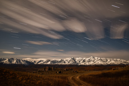 Colorful night landscape with clouds and stars in the form of tracks in the sky above snowy...