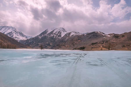 Picturesque spring view with a road through the snow and ice of a frozen mountain river with turquoise water against the backdrop of snowy mountains and a sky with clouds. Altai, Russiaの写真素材